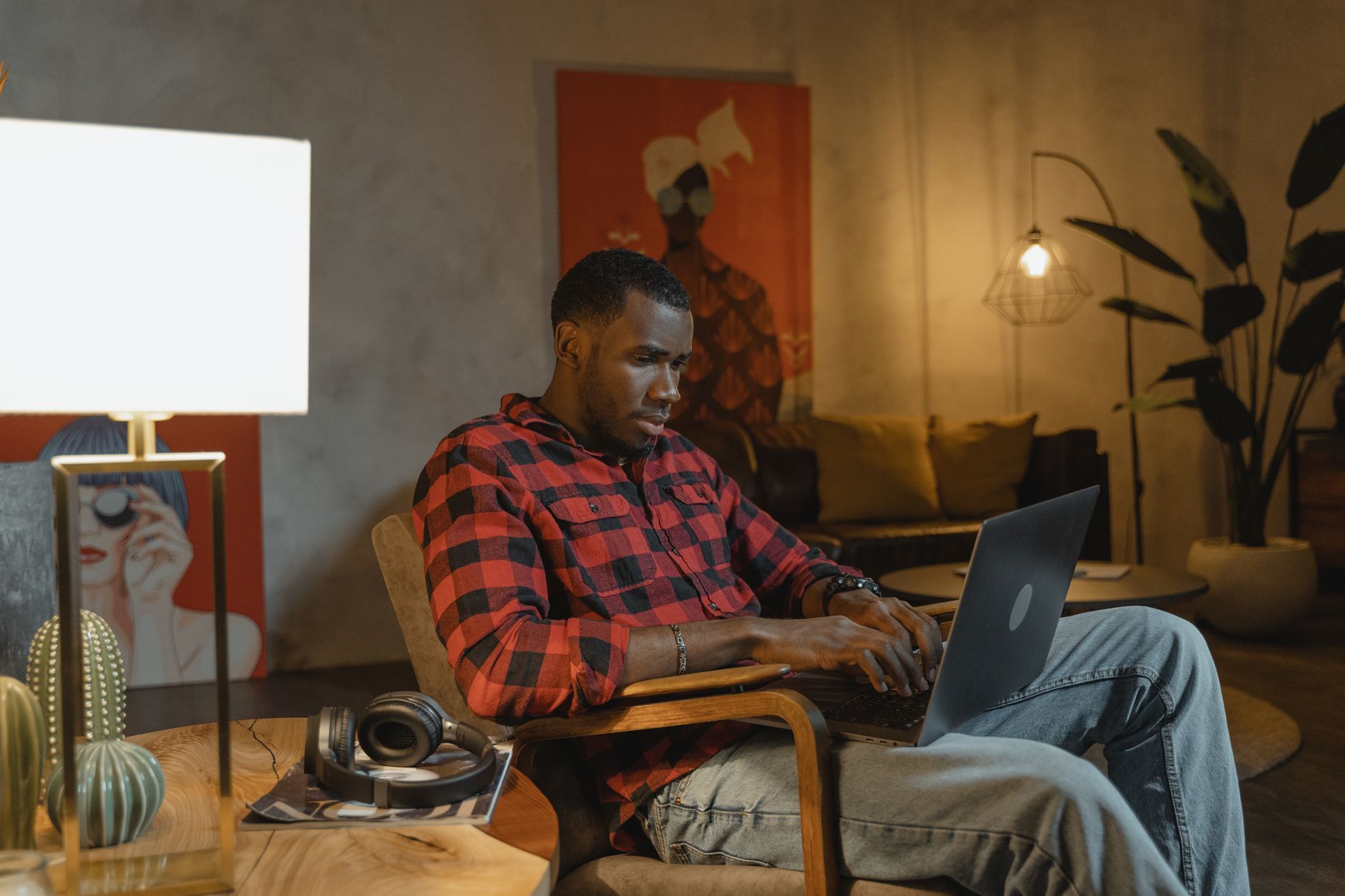 Un homme concentré tape sur un ordinateur portable, assis dans un fauteuil, dans un intérieur chaleureux avec des lampes et des plantes.
